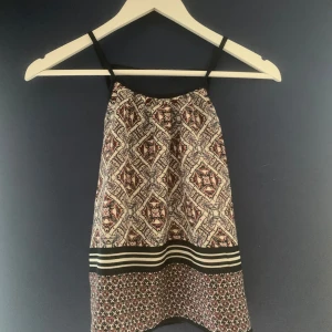 Mönstrad linne med smala axelband - Säljer ett snyggt linne med ett detaljerat mönster i olika färger. Toppen har smala axelband och en randig kantdetalj i svart och vitt längst ner. Perfekt till en varm sommardag. 