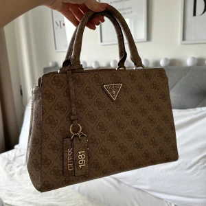 Brun handväska från Guess - Snygg brun handväska från Guess med monogrammönster och guldfärgade detaljer. Väskan har dubbla handtag och en praktisk dragkedja upptill. Inuti finns flera fack för enkel organisering. Designen på väskan går inte att få tag i längre!