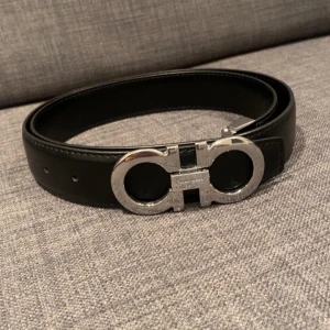 Svart skinnbälte med silverdetaljer - Snyggt svart skinnbälte från Salvatore Ferragamo med en iögonfallande silverfärgad spänne i form av två sammanlänkade cirklar. Perfekt för att ge en stilren touch till din outfit. Bältet har justerbara hål för bästa passform.