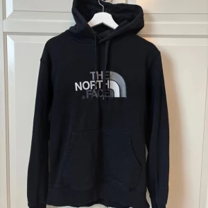 Svart hoodie från The North Face - Säljer en svart hoodie från The North Face med deras ikoniska logga i vitt och grått på bröstet. Tröjan har en klassisk känguruficka och justerbar huva med dragsko. Perfekt för en avslappnad stil.