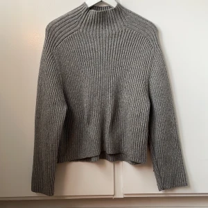 Grå ribbad stickad tröja - Säljer en stilren grå stickad tröja med ribbad struktur. Tröjan har långa ärmar och en hög hals, perfekt för kyligare dagar. Den är både bekväm och trendig, ett måste i garderoben!