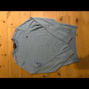 Ljusblå tröja från Morris - Säljer en stilren ljusblå tröja från Morris. Tröjan har långa ärmar och ett diskret emblem på bröstet. Perfekt för en avslappnad och snygg look.