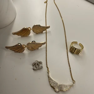 Smyckeset med guldiga vingar och ring - Elegant smyckeset med ett halsband och örhängen i form av guldiga vingar. Halsbandet har en kedja med en glittrig vingberlock. Setet inkluderar även en justerbar ring med nitar och en silverfärgad brosch.