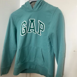 Grön hoodie från GAP - Säljer en snygg grön hoodie från GAP med klassisk logga på bröstet. Tröjan är knappt använd och i mycket bra skick. Säljer den pågrund av att den är för liten för mig. Tröjan har en känguruficka och en bekväm huva. Perfekt för en avslappnad stil. 