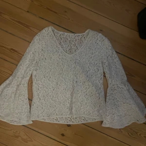 Vit spetsblus med vida ärmar - Säljer en elegant vit spetsblus med v-ringning och långa vida ärmar. Perfekt för en stilren look med vackert blommönster i spetsen. Passar bra till både jeans och kjol.