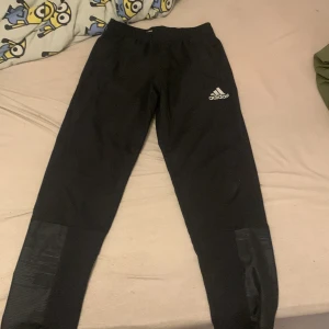 Svarta träningsbyxor från Adidas - Snygga svarta träningsbyxor från Adidas med elastisk midja och logga på benet. Perfekta för sport och träning. Byxorna har en modern design med detaljer vid benen.