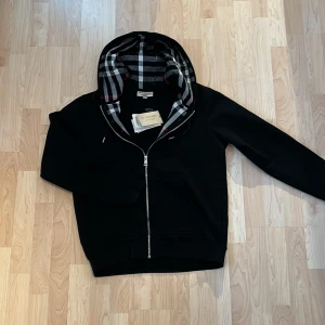 Svart huvtröja Burberry - Snygg svart huvtröja från Burberry med klassiskt rutigt mönster i huvan och broderad logga. Skriv gärna för fler bilder/info!