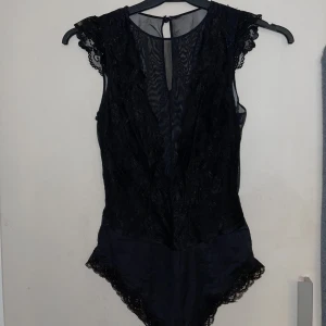 Svart spetsbody - Snygg svart body med spetsdetaljer och transparenta partier. Den har korta ärmar och en elegant design som passar perfekt för en kväll ute. Knytning i nacken för en säker passform.