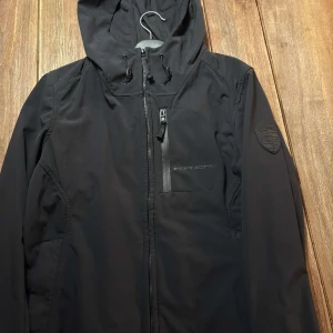 Svart jacka från Porsche - Snygg svart jacka från Porsche med huva och dragkedja framtill. Jackan har en diskret Porsche-logga på bröstet och ett märke på ärmen. Perfekt för en stilren look. En perfekt vindjacka! Kom privat vid funderingar!🤙