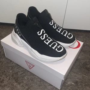 Svarta sneakers från Guess - Snygga svarta sneakers från Guess med vit sula och stort Guess-tryck på ovansidan. Skorna har en modern design med elastisk passform och är perfekta för en stilren look. Använda max 5 gånger. Skorna är i storlek 36 men hade även passat för dig med storlek 37