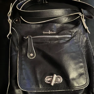 Svart axelväska från Gianni Conti - Snygg svart axelväska från Gianni Conti i skinn. Väskan har en praktisk dragkedja och ett stilrent spänne framtill. Perfekt för att bära med sig det nödvändigaste med stil.