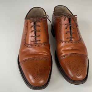 Läderskor från Loake 1880 - Eleganta bruna läderskor från Loake med klassiskt broguemönster. Modell;  ”strand brogue mahogany burnished calf”. Skorna har en låg klack, snörning och är tillverkade i England. Nypris 4200kr. 