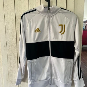 Vit och svart träningsjacka/fotbollströja Juventus, Adidas - Säljer en snygg vit och svart träningsjacka från Adidas med Juventus-logga i guld på bröstet. Jackan har dragkedja framtill och långa ärmar med svarta ränder längs axlarna. Perfekt för sportiga tillfällen eller som en stilren vardagsjacka. Tröjan har en liten fläck på baksidan men vet inte om den går bort i tvätten