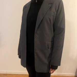 Grå kavaj från Bikbok - Snygg grå kavaj från Bikbok med klassisk design och två knappar framtill. Perfekt för en stilren look. Kavajen har långa ärmar och fickor med lock. Passar bra till både vardag och mer formella tillfällen.
