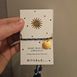 Blått armband från Rituals - Snyggt blått armband från Rituals med en guldmedaljong. Armbandet har en flätad design och en dekorativ tofs. Perfekt för att ge en touch av stil och elegans. Oanvänd, enbart öppnat förpackningen för bild 🤗