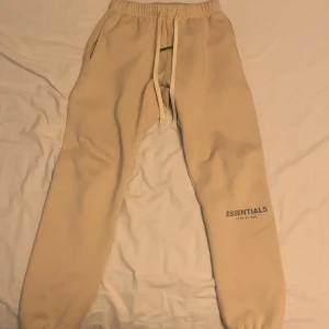 Beige mjukisbyxor från Essentials - Snygga beige mjukisbyxor från Essentials Fear of God med elastisk midja och snörning. Byxorna har en avslappnad passform och är perfekta för en bekväm stil. Essentials-loggan är tryckt på benet.