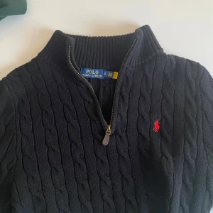 Svart stickad tröja från Ralph Lauren - Säljer en stilren svart stickad tröja från Ralph Lauren med kabelstickat mönster och dragkedja vid halsen. Tröjan har en liten röd logotyp på bröstet och är i ett mycket gott skick! Storleken är S men den sitter mer som M. Skriv vid frågor!