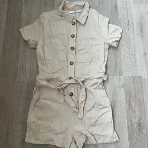 Beige jumpsuit med knappar och knytband - Snygg beige jumpsuit med korta ärmar och knappar framtill och ett knytband i midjan. Perfekt för en avslappnad stil.