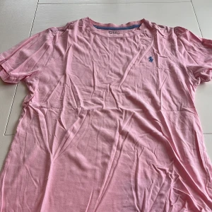 Rosa t-shirt från Ralph Lauren - Säljer en snygg rosa t-shirt från Ralph Lauren med det klassiska broderade logotypen i blått på bröstet. T-shirten är kortärmad och tillverkad i mjuk bomull. Perfekt för en avslappnad stil. 🌸