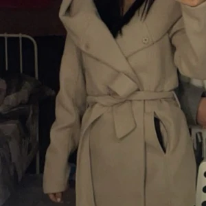 Beige kappa med bälte - Snygg beige kappa med bälte och stora slag. Perfekt för kyligare dagar med sin långa design och knappar framtill. En stilren och tidlös jacka som passar till många tillfällen.