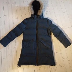 Mörblå jacka från Geographical Norway - Säljer en svart dunjacka från Geographical Norway med pälskantad huva och dragkedja framtill. Jackan har långa ärmar och praktiska fickor med knappar. Perfekt för kalla vinterdagar, jackan har lite skavanker vid änden på ärmarna och lite på dragkedjan men har annars ett fint helhetsintryck