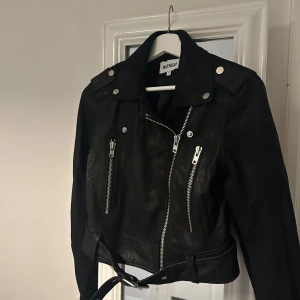 Svart skinnjacka från Weekday - Snygg svart skinnjacka från Weekday med dragkedja och bälte i midjan. Jackan har långa ärmar och silverfärgade detaljer. Perfekt för en cool look!