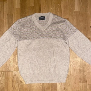 Beige stickad tröja med mönster - Säljer en beige stickad tröja från Glenmore med ett diskret mönster i grått och vitt över bröstet. Tröjan har en v-ringad hals och långa ärmar, perfekt för kyligare dagar. Den är både stilren och bekväm.