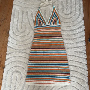 Randig klänning från Gina Tricot - Snygg randig klänning från Gina Tricot i härliga färger som orange, blå, turkos och vit. Klänningen har en v-ringad design med knytband i nacken och är perfekt för sommarens äventyr. Den är stickad och har en kort längd som ger en avslappnad och trendig look.