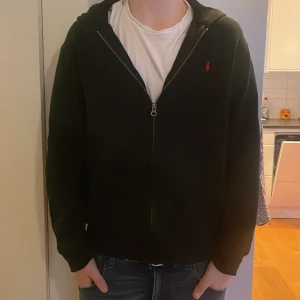 Svart hoodie från Polo Ralph Lauren - Säljer en svart hoodie från Polo Ralph Lauren med dragkedja och en liten röd logga på bröstet. Perfekt för en casual look. Den har långa ärmar och en bekväm passform. Hör av er vid funderingar!