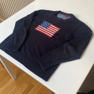 Stickad Tröja me flagga - Säljer en mörkblå stickad tröja från GANT med en broderad amerikansk flagga på bröstet. Tröjan har långa ärmar och ribbade muddar vid ärmslut och nederkant. Perfekt för en stilren och avslappnad look.