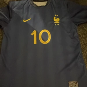 Blå fotbollströja från Nike - Säljer en blå fotbollströja från Nike med nummer 10 och namntryck på ryggen. Tröjan har korta ärmar och ett broderat emblem på bröstet. Perfekt för fotbollsfans!