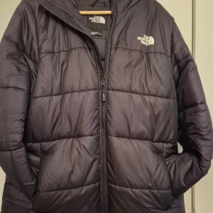 Svart dunjacka från The North Face - Säljer en svart dunjacka från The North Face med dragkedja och huva. Jackan har en quiltad design och är perfekt för kyliga dagar. Den har två sidofickor och logotypen på bröstet. Perfekt för vintern! 🖤