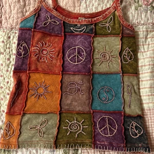 Färgglatt linne med broderade symboler - Unik linne med broderade symboler i olika färger som orange, grön, lila och blå. Toppen har smala axelband och en patchwork-design med olika symboler som sol och fredstecken. Perfekt för en bohemisk eller hippiestil! Gjort av ganska elastiskt och mjukt tyg! Sitter lite större på mig som S