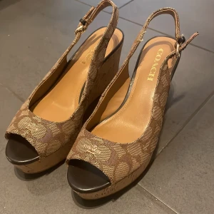 Bruna kilklackar från Coach - Snygga bruna kilklackar från Coach med öppen tå och justerbar rem runt hälen. Skorna har ett mönster i olika bruna nyanser och en korkliknande sula som ger en somrig känsla. 