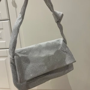 Silver axelväska med glittriga detaljer - Snygg silver axelväska med glittriga detaljer och en vriden axelrem. Väskan har en praktisk dragkedja inuti för säker förvaring. Perfekt för att lägga till lite bling till din outfit!