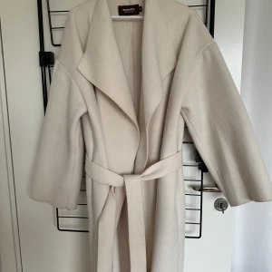 Beige kappa Binacas collection med Nelly  - Elegant beige kappa från Nelly med knytbälte i midjan. Den har en klassisk design med långa ärmar och breda slag. Perfekt för en stilren look.