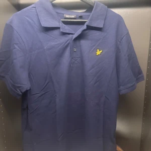 Blå pikétröja från Lyle & Scott - Säljer en stilren blå pikétröja från Lyle & Scott med klassisk krage och knappslå. Tröjan har en broderad gul logotyp på bröstet. Perfekt för en avslappnad men ändå snygg look.