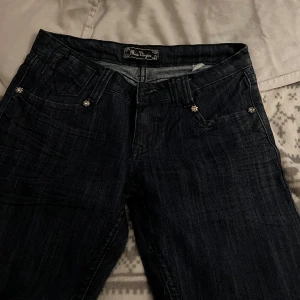 Mörkblå jeans med strassdetaljer - Snygga mörkblå jeans från Miss Me med strassdetaljer på bakfickorna. Jeansen har en klassisk femficksdesign och en lågmidjad passform. Perfekta för en stilren look med en touch av glamour.