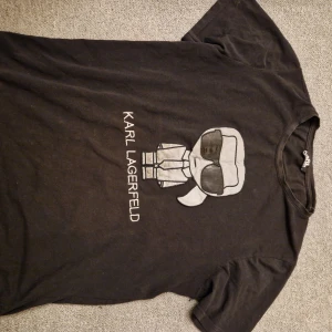 Svart t-shirt med Karl Lagerfeld-tryck - Snygg svart t-shirt med ett coolt tryck av Karl Lagerfeld i grått och vitt. Perfekt för dig som gillar mode och vill ha en stilren look. T-shirten har korta ärmar och en klassisk passform.