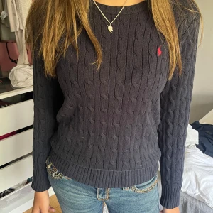 Mörkblå stickad tröja från Ralph Lauren - Säljer en snygg mörkblå stickad tröja från Ralph Lauren. Den har ett klassiskt kabelstickat mönster och en liten röd logga på bröstet. Tröjan är långärmad och har ribbade muddar vid ärmslut och nederkant. Perfekt för höst och vinter!