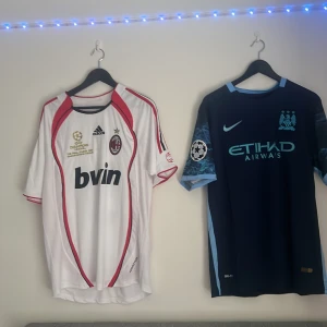 Fotbollströjor från AC Milan och Manchester City - Säljer två fotbollströjor. Den första är en vit AC Milan-tröja med röda och svarta detaljer, kortärmad och med 'Maldini 3' på ryggen. Den har Champions League-brodyr och är från Adidas. Den andra är en mörkblå Manchester City-tröja med ljusblå detaljer, kortärmad och med 'Kun Agüero 10' på ryggen. Den är från Nike och har Etihad Airways som sponsor.