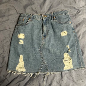 Jeanskjol med slitningar - Säljer en cool jeanskjol med slitningar och fransig kant. Perfekt för en avslappnad och trendig look. Passar till allt och bra att ha. Kjolen har två fickor bak och en klassisk jeansknäppning fram. Aldrig använt. Kan klippas till önskad minikjol.