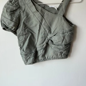 Grön oneshoulder topp från Zara - Säljer en snygg grön oneshoulder topp från Zara. Den har en cool knytdetalj framtill och puffiga korta ärmar. Perfekt för sommarens fester eller en dag på stan. Toppen är i bra skick och superbekväm att ha på sig!