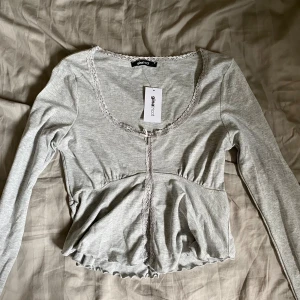 Grå långärmad topp från Gina Tricot - Säljer en helt ny superfin grå långärmad topp från Gina Tricot. Den har en söt spetsdetalj längs halsringningen och knäppning framtill. Toppen är lite croppad och har en volangkant nedtill. Perfekt för både vardag och fest!