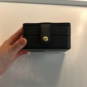 Svart smyckeskrin med spegel - Praktiskt svart smyckeskrin med en spegel på insidan av locket. Skrinet har flera fack för förvaring av smycken, inklusive ringhållare och krokar för örhängen. Perfekt för att organisera dina smycken hemma eller på resan.