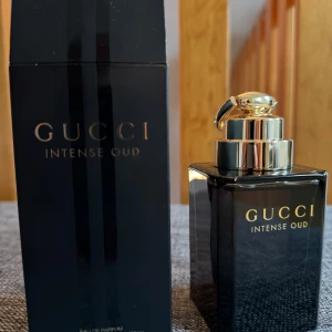 Gucci Intense Oud Parfym - Elegant parfymflaska från Gucci i svart med guldlock. Doften Intense Oud är känd för sina rika och djupa noter, perfekt för den som älskar en intensiv och lyxig doftupplevelse. Flaskan har en stilren design med märkets logotyp i guld på framsidan. Endast testad. Flaskan kommer i 90ml storlek.