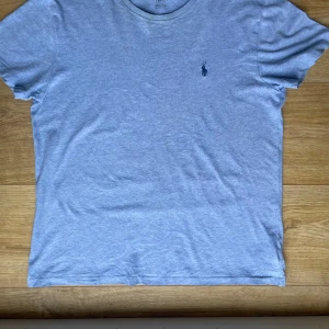Grå t-shirt från Polo Ralph Lauren - Säljer en stilren grå t-shirt från Polo Ralph Lauren med det klassiska broderade logotypen på bröstet. T-shirten är kortärmad och tillverkad i mjuk bomull för en bekväm passform. Perfekt för en avslappnad look.
