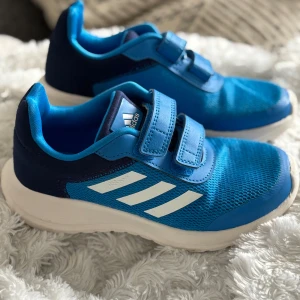 Blå sneakers från Adidas - Säljer ett par blå sneakers från Adidas med vita detaljer och kardborreband. Skorna har en sportig design med meshmaterial för bra ventilation. Perfekta för en aktiv livsstil. Normal passform. 