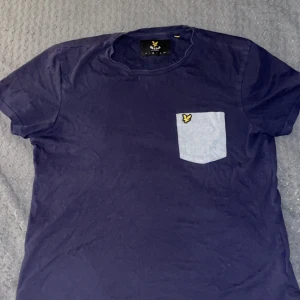 Mörkblå Lyle & Scott tröja - Säljer en mörkblå t-shirt från Lyle & Scott med en ljusblå bröstficka och deras ikoniska logga. Perfekt för en avslappnad stil. T-shirten är kortärmad och gjord i mjukt material.