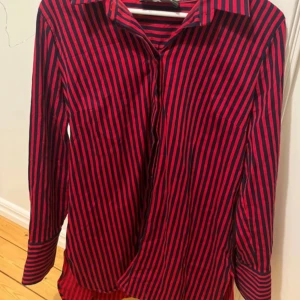 Röd och svart randig skjorta från By Malene Birger - Snygg röd och svart randig skjorta från By Malene Birger. Skjortan har långa ärmar och klassisk krage, perfekt för en stilren look. Tillverkad i en bomullsblandning för en bekväm passform. Liten i storleken så skulle säga att den passar M/38. Säljer en likadan i storlek M/38 och passar precis som en M/38.